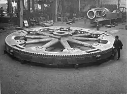 Flywheel for a large rolling mill engine 1900; the heavy rim is cast in four sections bolted together at the rim. Top right, the trunk guide and bedplate of the engine under manufacture, beyond the bedplate is the flywheel and connecting rod of a small horizontal steam engine.[80][120]