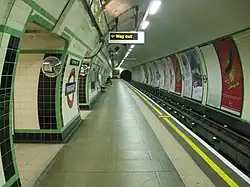 View along a platform in a circular tunnel. Tracks run on the right side with the walls covered with cream and green tiles; an illuminated 'way out' sign overhead indicates the exit.