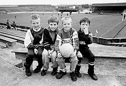Four young boys sit on concrete steps at a football ground, one holding a football, with a pitch and stands visible in the background.