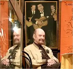 Horacio Ferrer wearing a brown and white polka dot shirt and tan jacket, sitting in a chair with hands crossed on his lap, with a painting of three male guitarists behind him