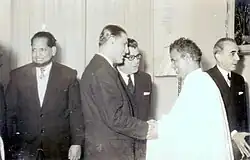 A smiling Babana, in white, greets men in suits.
