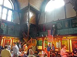 A large space with a wooden gallery along the upper portion, large windows above, and paintings and ornate decorations in the wooden walls. On the lower level, at the bottom of the image, there are a lot of people doing various things
