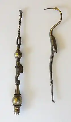 Two long, thin metal objects. The right object resembles a double-ended dentist pick in the shape of a crane bird. Its legs form the lower half, ending in a small pick; the body and wings form the handle in the middle; whereas the curved neck, head and long pointed beak form the upper pick. The left object is straight; a thin twisted rod ends in a large ball, followed by a fish motif again bounded by a ball, and ending in a crown with its peaks pointing inwards, presumably to hold bristles.
