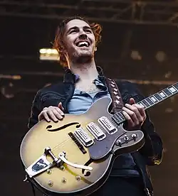 A singer, clothed in a white shirt and a brown jacket, is holding a guitar and singing