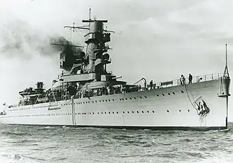 A black-and-white photograph of the cruiser in a calm sea, seen from the bow. Her entire profile is visible, which includes a tall, dominant tower, a large turret on the deck, a smaller turret superimposed near the bridge, and a funnel releasing smoke.