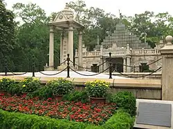 A well-maintained memorial site features a large monument with stone pillars and a triangular pediment inscribed with Chinese characters. The foreground displays vibrant flower beds with red and yellow blossoms, surrounded by green bushes and ornamental plants. The monument is fenced off by a chain and stanchion barrier. In the background, a multi-tiered stone structure with more Chinese inscriptions and decorative stonework is visible, flanked by trees.