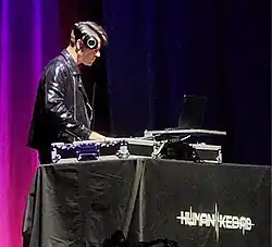 Jason Parsons, aka Human Kebab, seen from right profile, wearing a black leather jacket, large headphones perched on the side of his head, operating a control board onstage
