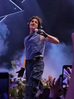 Male singer performing onstage, wearing headphones, a sleeveless blue vest over a white shirt, black gloves, and cargo pants, holding a microphone toward the audience.