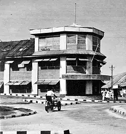 Intersection, not busy, black and white, the old days. Dude out front cruisin on an old motocycle. Couple of teenage women chillin out front. Building in the center of frame, roundish, squarish, three floors visible including ground floor.