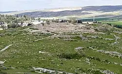 View of Horvat Rimmon on a low hill, with stone ruins spread across the summit and terraces, surrounded by green rocky slopes, with a modern village and rolling hills in the background