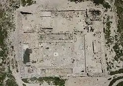 Aerial view of the synagogue at Horvat Rimmon, showing a rectangular paved hall with a small central panel, surrounded by low stone walls and adjoining rooms amid sparse vegetation on a hilltop