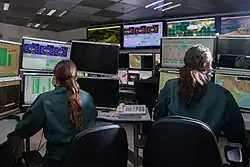 A view from behind of two satellite operators in Unit 9900, sat in front of many displays etc.