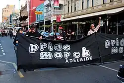People on street holding a black sign with silver lettering that says "Pride March Adelaide".