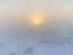 A shot of the sun hanging low over some short buildings. The sun is reddish, and the entire shot is heavily obscured by fog.