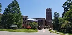 A red brick building is seen, with grass, trees, and construction equipment in the foreground. The building's tower and main entrance are to the right, with glass roofs and windows stretching to the left. Architecturally, the tower echoes the shape of the tower in the original building.