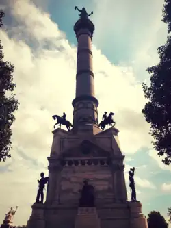 The Iowa Civil War Monument shown at an angle. The monument has a base with two statues at the bottom of the base, and two more statues of people on horses at the top of the base. In the middle of the base, there is a pillar, and on top of the pillar is another statue of a man.
