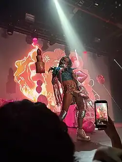 Isabella Lovestory at her 2025 Vanity tour, performing at her concert in the Brooklyn venue Elsewhere. She holds her microphone with a giant glittering tarantula with her right hand while her left hand puts an oversized fake glittery lipstick to her crotch.