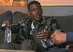 Rogers in a couch facing slightly left, head tilted slightly, gesticulating with left hand while holding an unlabeled disposable plastic water bottle in the right hand.
