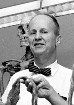 Balding man in a striped bowtie standing in front of a skeleton