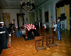 casket draped with a flag in a large room. Several soldiers stand around it, and two clerics kneel on the kneelers at the side. Two empty kneelers are in the foreground