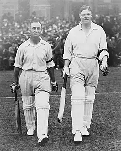 Two batsmen walk on to a cricket ground with a crowd behind them