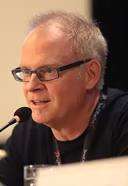 Portrait of an older man with pattern hair loss wearing glasses and a black t-shirt speaks into a lectern microphone.