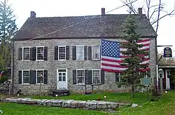 A large stone house with a large American flag draped across its upper right story behind an evergreen tree. In front of it is a blue and gold historical marker.