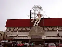 A two-storeys building with a red roof. On the façade and the roof, a figure of a moving man holding a long basket on his right hand and a helmet on his head. Since it is day, the neon tubes shaped as different phases of the player movement are barely visible. Over the figure, the letters "Ja[missing i] Alai". Cars are parked before the building.