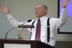Spann in a suit and suspenders in front of a podium, raising his hands above his head