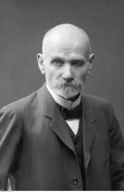 An older bald man with a large mustache and short beard in a suit with a bowtie