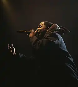 A black man, dressed in all black, stands side on to the camera. He is performing on stage with a microphone in his hand