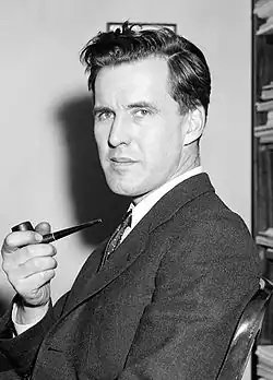 A young man wearing a suit and tie turns profile to the camera, holding a pipe in his hand.