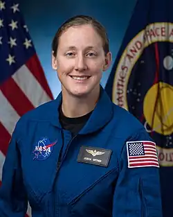 A woman in a blue flight suit is sitting in front of US & NASA flags; she is facing the camera and smiling.