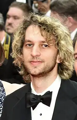 Johan Lenox, a mid-thirties white male with blonde neck-length curls, noticeably lighter on top. He has dark eyes and eyebrows and a short dark beard, though his moustache appears lighter, closer to his hair color. He looks directly at the camera with a neutral expression, dressed in formal attire with a black bow tie. In the blurred background, photographers are lined up at a film premiere, with two other figures faintly visible on either side of him.