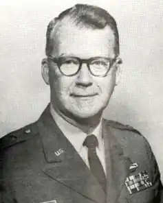 Black and white portrait of a caucasian male, short dark hair, dark rimmed glasses, clean shaven; wearing a light colored shirt, dark tie, dark jacket. Jacket has US emblem on left lapel; military ribbons on right chest
