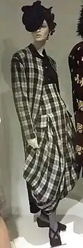 A mannequin is dressed in a black and white plaid ensemble with an oversized draped skirt, matching jacket, dark blouse, and a large asymmetrical black hat