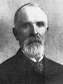 Black-and-white photograph of a man with thin hair and a beard in a suit and tie, with a stern expression