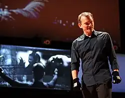 A man wearing a gray shirt, brown pants and black 3D tracking gloves stands in front of a screen.