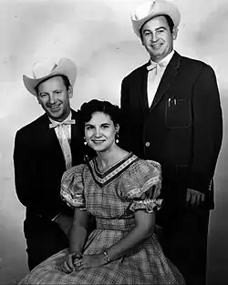 Two men wearing dark suits and cowboy hats standing either side of a woman wearing a gingham dress