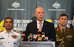 Dutton at a press conference with two men in military uniforms. They are standing in front of a black backdrop with white text reading "ZERO CHANCE OF ILLEGAL MIGRATION".