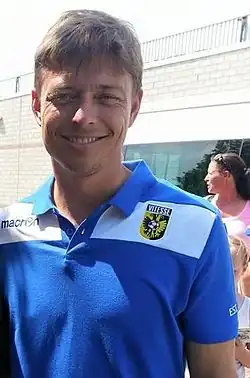 A photograph of a man in an external setting with two people partly covered visible in the background. The man is wearing a blue shirt with white details.