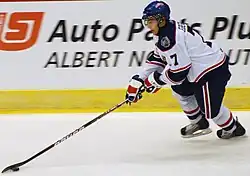 An ice hockey player in his late teens in mid-stride, carrying the puck. He is looking down towards the puck on his outstretched stick.