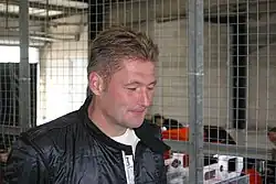 Photo of Jos Verstappen, in a leather jacket, posing in front of a fence