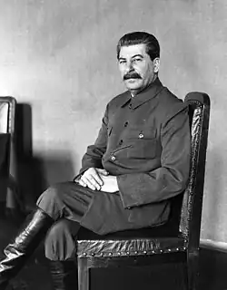 Man with a mustache wearing a buttoned military jacket and tall boots sitting on a wooden chair with hands folded