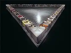 A large, triangular dining table with ornately decorated plates and tableclothes in a dark room. The space encapsulated between the three sides of the table shows a floor with the written names of famous women.