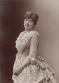 Sepia-toned portrait of a white woman, standing; her hair is dressed in curls atop her head; she is wearing a dress with calico-print skirt and front panel.