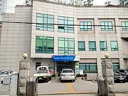 A three storey tall grey building with a blue sign above the door, which has Korean text written on it