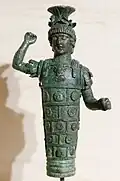 Photograph of an ancient bronze statuette of a made god with ornate armor, raised arm, and helmet, evoking strength and historical significance against a neutral background.