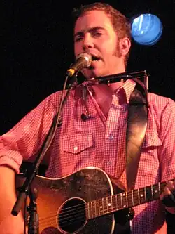 blurry image of Justin Rutledge wearing a light-coloured checkered shirt, playing acoustic guitar and singing into a microphone onstage