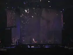 A stage with a big screen display showing a man with a beard. Sitting in a piano, a man with long curly hair wears a white shirt, with the sleeves roll back, and a black vest, plays the piano. To the man's left, a man can be seen standing playing an electric piano.